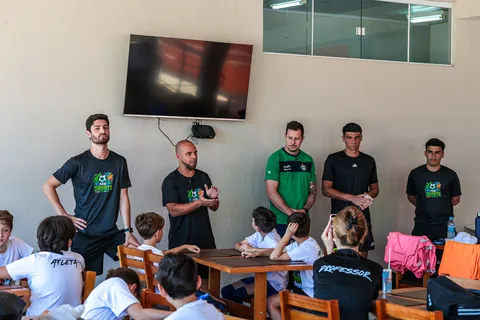 Professores de camiseta preta e verde em pé, com a turma sentada às mesas