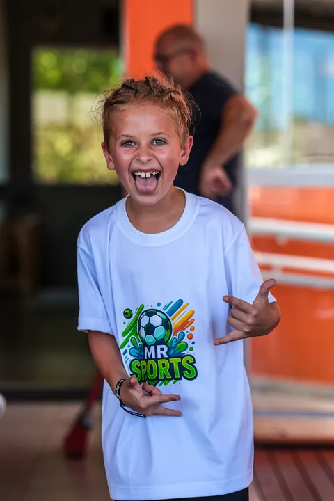 Menino de camiseta branca da MR Sports sorri e faz sinal com as mãos