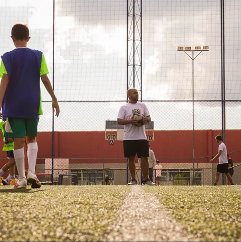 Professor segura a bola no meio do campo enquanto as crianças se posicionam