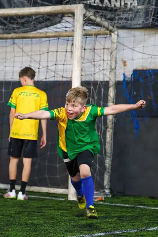 Menino de colete verde comemora com os braços abertos em frente ao gol