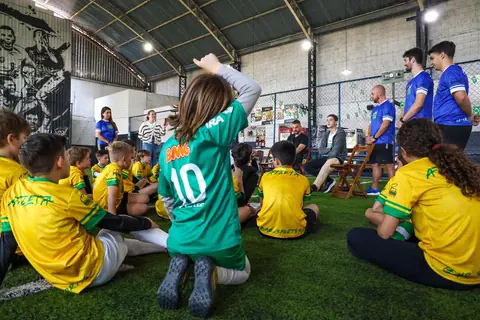 Crianças sentadas no campo ouvem os convidados, uma delas de pé com a camisa 10