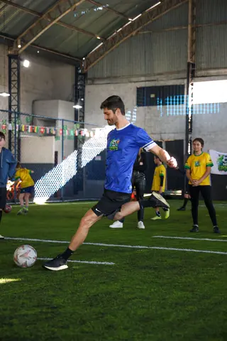 Professor de camiseta azul da MR Sports conduz a bola no meio das crianças